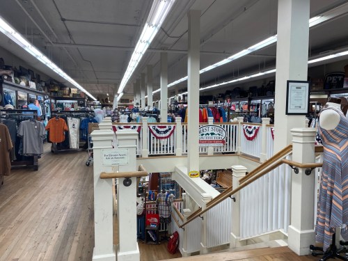 Interior of a retail store with clothing displays, wooden floors, and patriotic decorations. Stairs lead to a lower level.