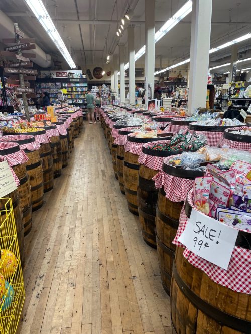 A grocery store aisle lined with wooden barrels filled with snacks, featuring a sale sign and a shopper in the background.