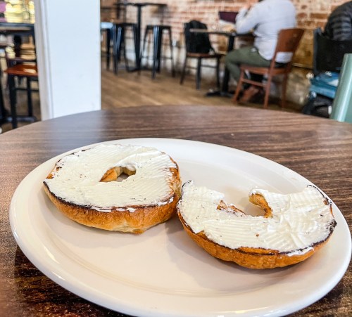 A toasted bagel with cream cheese on a white plate, set on a wooden table in a café.