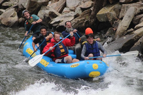 A group of five people in a blue raft navigating a rocky river, smiling and paddling through white water.