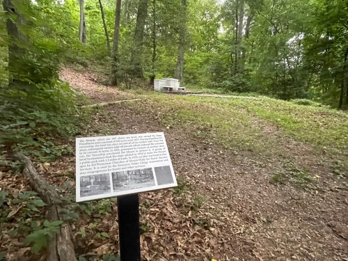 A sign in a wooded area describes historical significance, with a stone structure visible in the background.