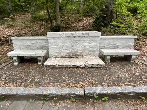A stone bench with the words "Holy Holy Holy" carved on it, surrounded by trees and fallen leaves.