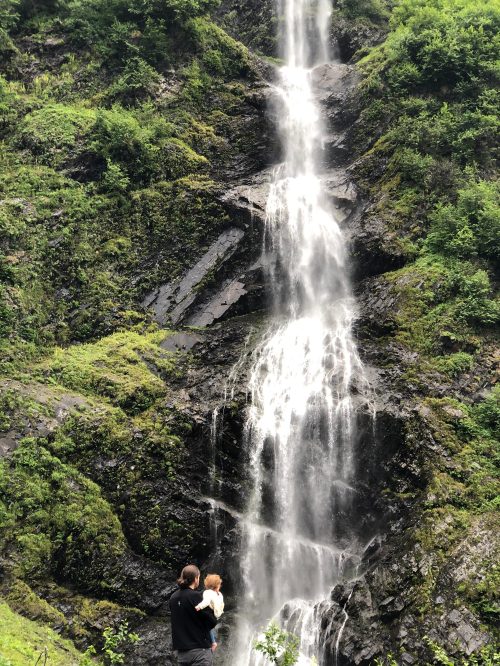 A person holds a child while standing in front of a tall waterfall surrounded by lush greenery.