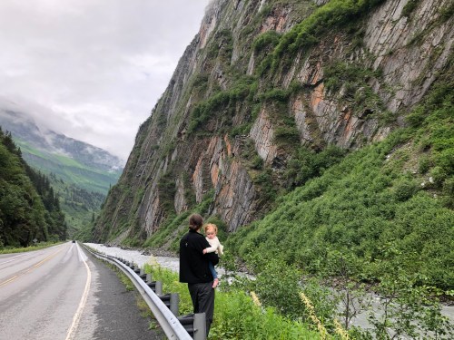 A person holds a child while standing by a scenic road, surrounded by steep, lush green cliffs and a river.