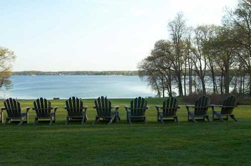 A row of wooden chairs facing a serene lake, surrounded by trees and a clear sky.