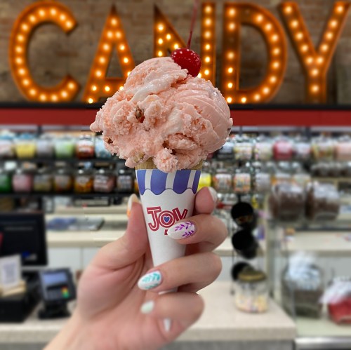 A hand holds a cone of pink ice cream topped with a cherry, with a candy shop backdrop.
