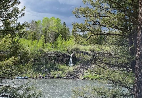 A serene river scene with a waterfall in the distance, surrounded by lush greenery and trees under a cloudy sky.