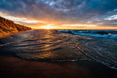Sunset over a beach with gentle waves, reflecting golden light on the water and dramatic clouds in the sky.