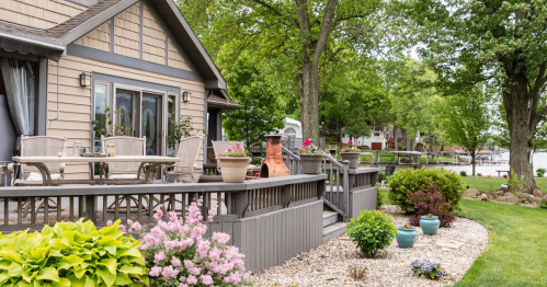 A cozy lakeside home with a deck, surrounded by greenery and colorful flowers, overlooking the water.