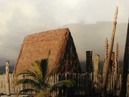 A traditional thatched hut surrounded by wooden totems and a palm tree, set against a cloudy sky.