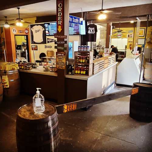 A rustic food counter with barrels, hand sanitizer, and signs promoting social distancing in a casual dining space.