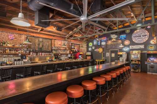 A vibrant bar interior with a long counter, colorful neon signs, and vintage arcade games in a rustic setting.