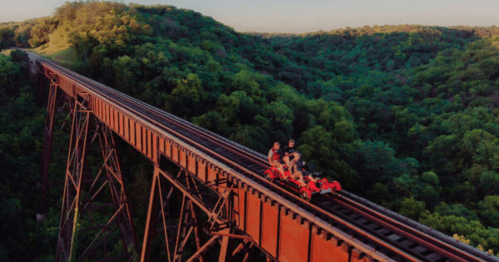 Two people ride a red rail bike on an elevated train track surrounded by lush green hills.