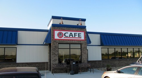 Exterior of a cafe building with a blue roof and large sign reading "The Cafe." Clear sky in the background.