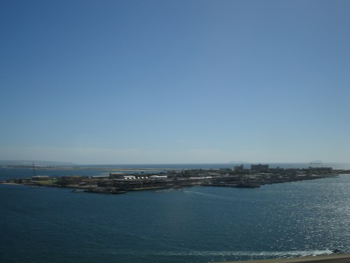 A coastal view of an island with buildings, surrounded by blue water under a clear sky.
