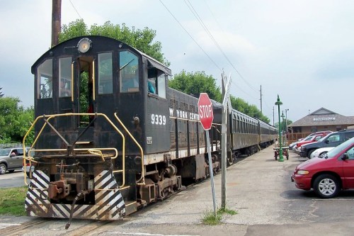 A vintage black train engine parked near a stop sign, with passenger cars and parked cars in the foreground.