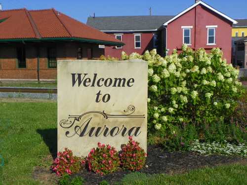 A stone sign reading "Welcome to Aurora" surrounded by colorful flowers and greenery, with buildings in the background.