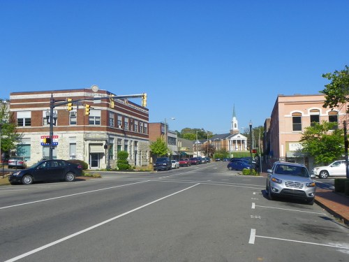A quiet downtown street with brick buildings, parked cars, and a church steeple in the background under a clear blue sky.