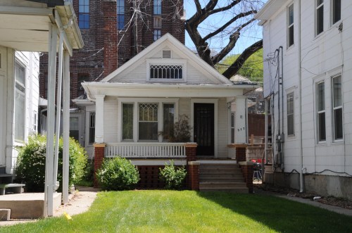 A small, charming house with a front porch, surrounded by greenery and adjacent to taller buildings.
