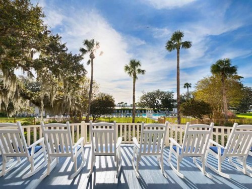 A row of white chairs on a deck overlooking a lush landscape with palm trees and a blue sky.