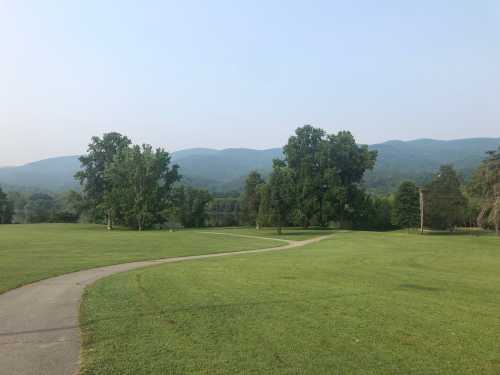 A winding path through a grassy park with trees and mountains in the background under a hazy sky.