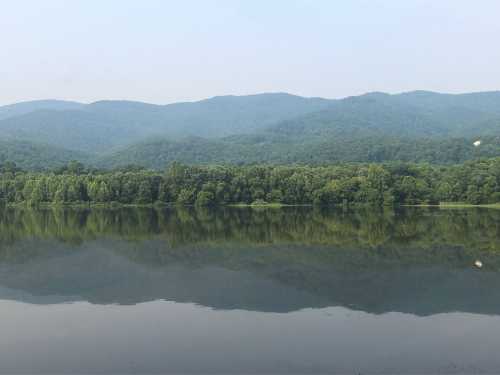 A serene lake reflecting green mountains under a hazy sky, surrounded by lush trees.