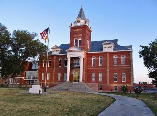 Historic red brick building with a clock tower, surrounded by trees and a grassy area, under a clear blue sky.
