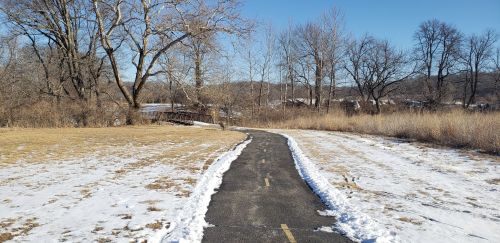 A snow-covered path winds through a winter landscape with bare trees and grassy areas.