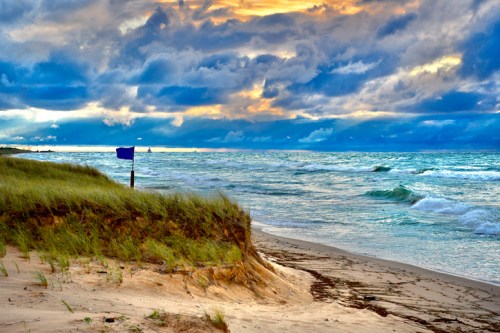 A serene beach scene with rolling waves, grassy dunes, and a dramatic sky filled with clouds at sunset.
