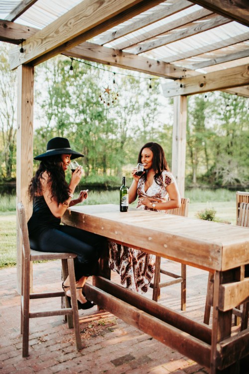 Two women sit at a wooden table outdoors, enjoying drinks and chatting in a lush, green setting.