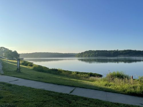A calm lake at sunrise, surrounded by greenery and a clear blue sky, with a path along the water's edge.