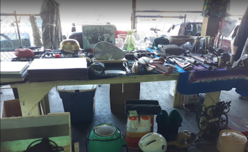 A cluttered table filled with various items, including tools, kitchenware, and collectibles, in a market setting.