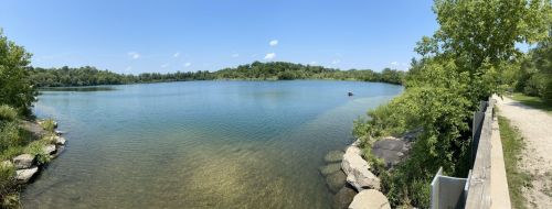 A serene lake surrounded by greenery under a clear blue sky, with a path along the water's edge.