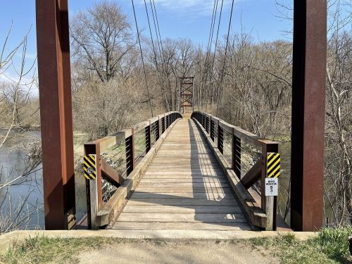 A wooden suspension bridge spans a calm river, surrounded by bare trees and a clear blue sky.