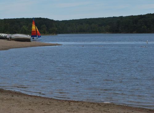 A calm lake scene with a colorful sailboat on the shore, surrounded by trees and sandy beach.