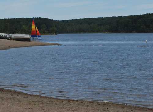 A calm lake scene with a colorful sailboat on the shore, surrounded by trees and sandy beach.