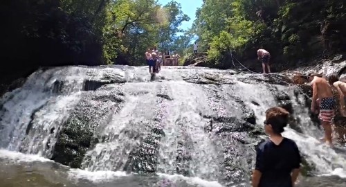 A group of people enjoying a sunny day at a waterfall, with some climbing and others playing in the water.