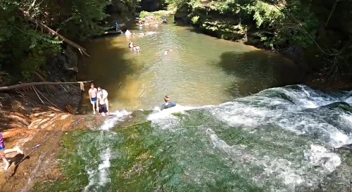 A scenic waterfall flows into a calm pool, with people swimming and enjoying the natural surroundings.