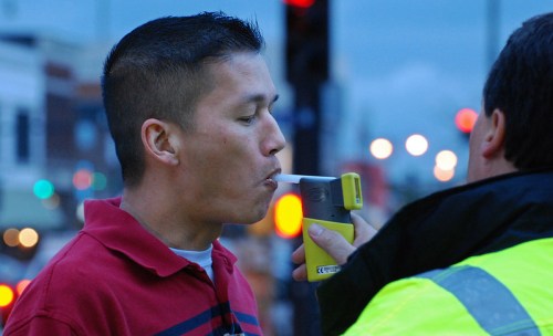 A man blows into a breathalyzer while a police officer observes, with city lights blurred in the background.