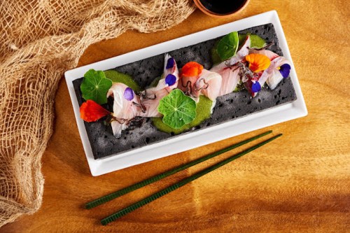 A beautifully arranged dish of sashimi on a black plate, garnished with edible flowers and green sauce, with chopsticks nearby.