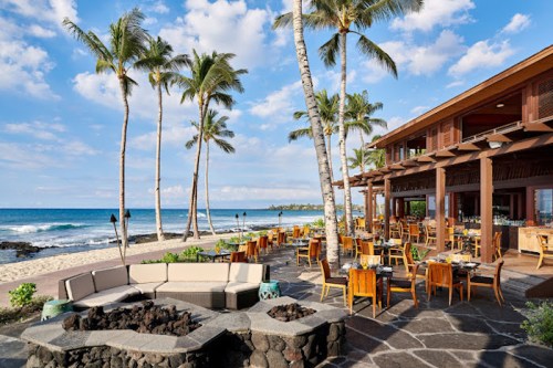 A beachfront restaurant with palm trees, outdoor seating, and a fire pit, overlooking the ocean and blue sky.