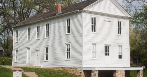 A historic white two-story house with multiple windows, set on a stone foundation surrounded by greenery.