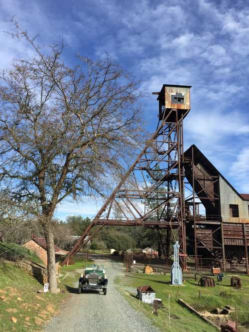 An old mining structure with a tall tower, surrounded by trees and vintage machinery on a gravel path under a blue sky.