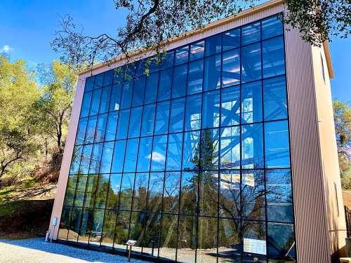 Modern building with large glass windows reflecting trees and blue sky, surrounded by greenery.