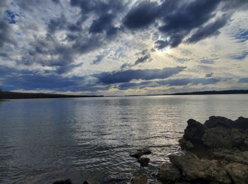 A serene seascape with calm waters, rocky shore, and dramatic clouds reflecting sunlight in the sky.