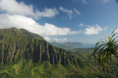 Lush green mountains under a blue sky with clouds, overlooking a coastal landscape and distant hills.