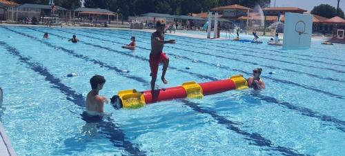 Children playing on a floating balance beam in a swimming pool, with others swimming nearby on a sunny day.