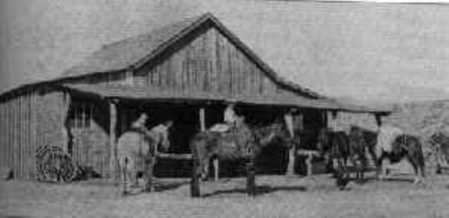 Black and white image of a barn with horses and people standing outside.