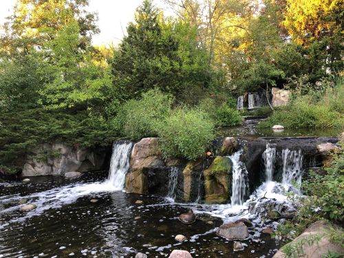 A serene waterfall cascades over rocks, surrounded by lush greenery and trees, reflecting warm sunlight.