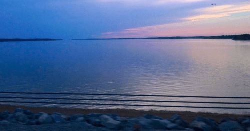 A serene lake at sunset, with calm waters reflecting shades of blue and pink, framed by a rocky shoreline.